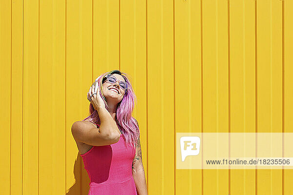 Happy young woman enjoying music listening through wireless headphones in front of yellow wall
