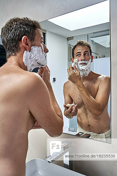 Man looking at mirror reflection and shaving in bathroom