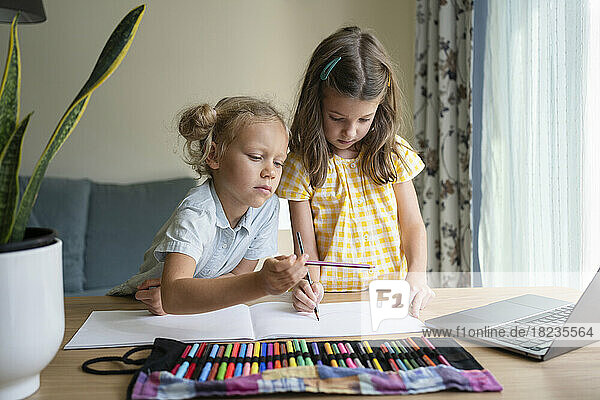 Mädchen zeichnen gemeinsam auf einem Skizzenblock am Laptop am Schreibtisch