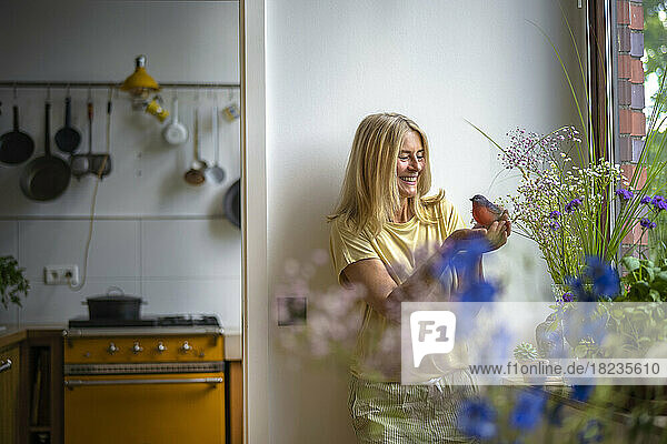 Happy mature woman holding bird standing in front of wall at home