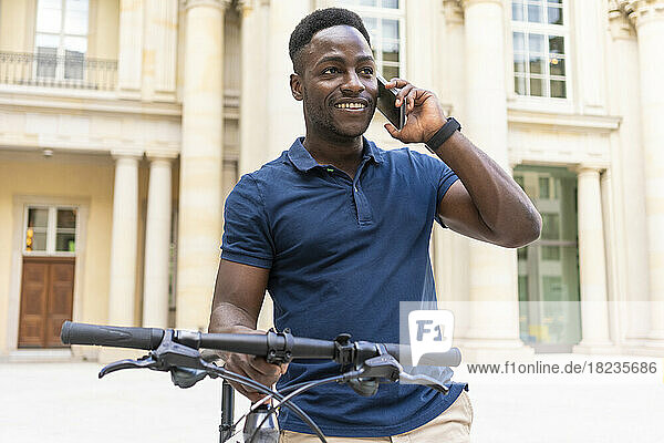 Smiling man talking on phone standing with bicycle in front of building