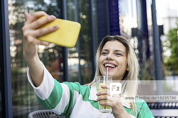 Glückliche junge Frau hält ein Trinkglas in der Hand und macht ein Selfie mit dem Mobiltelefon