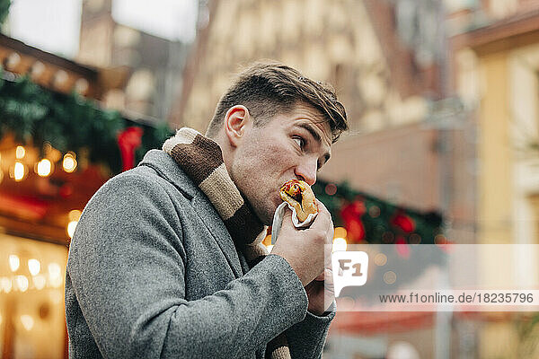 Junger Mann isst Hot Dog und steht auf dem Weihnachtsmarkt