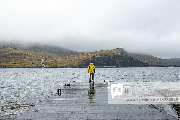 Man standing on pier in front of sea and mountains under cloudy sky