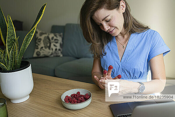 Smiling businesswoman putting raspberries on fingers at desk in home office