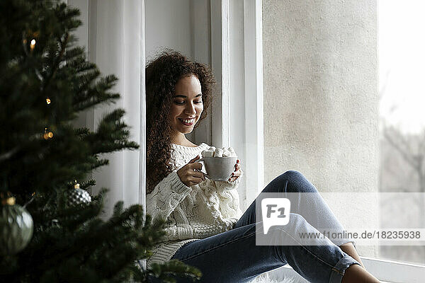 Happy young woman sitting on window sill with cup of marshmallow cocoa at home