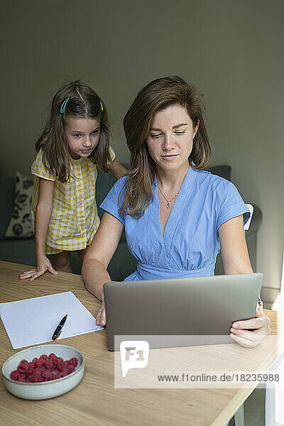 Freelancer working on laptop with daughter standing by desk at home