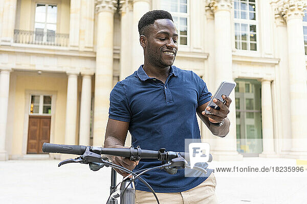 Smiling man using smart phone holding bicycle in front of building