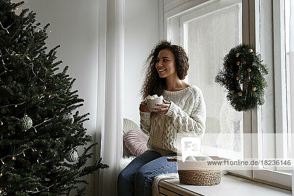 Happy woman sitting on window sill with cup of marshmallow cocoa at home
