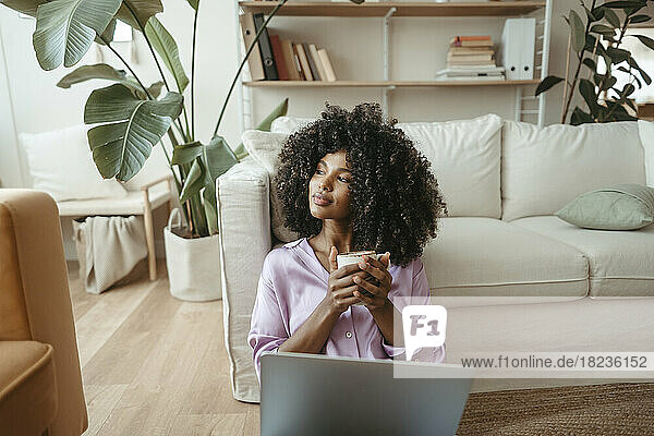 Nachdenkliche Frau mit Kaffeetasse und Laptop im Wohnzimmer