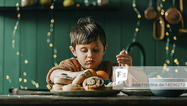 Boy holding sieve in front of cinnamon buns and oranges on table