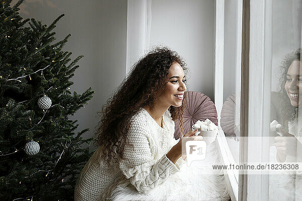 Happy young woman with cup of marshmallow cocoa looking out through window