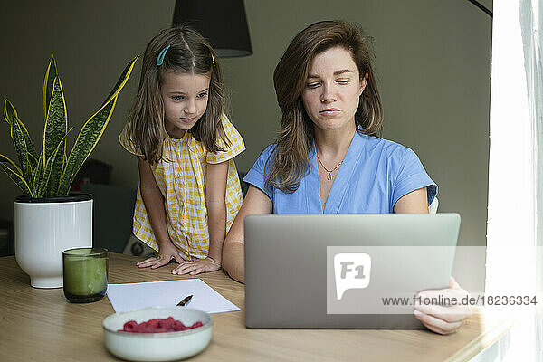 Businesswoman working on laptop with daughter standing by desk at home