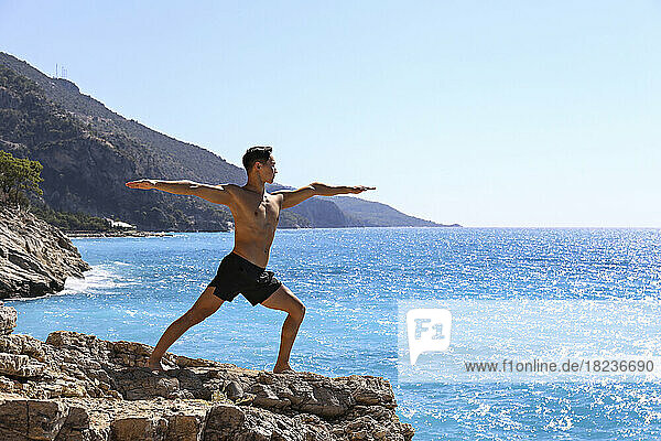Shirtless man practicing Virabhadrasana yoga pose standing on rock near sea