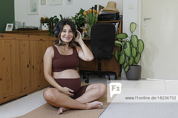 Glückliche schwangere Frau  die zu Hause auf einer Yogamatte sitzt und Musik über drahtlose Kopfhörer hört