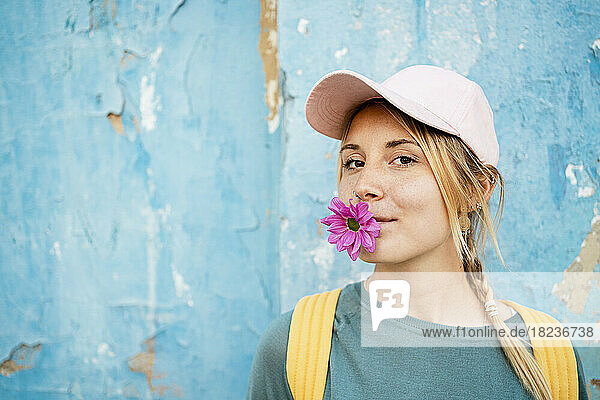 Young woman wearing cap holding pink flower in mouth