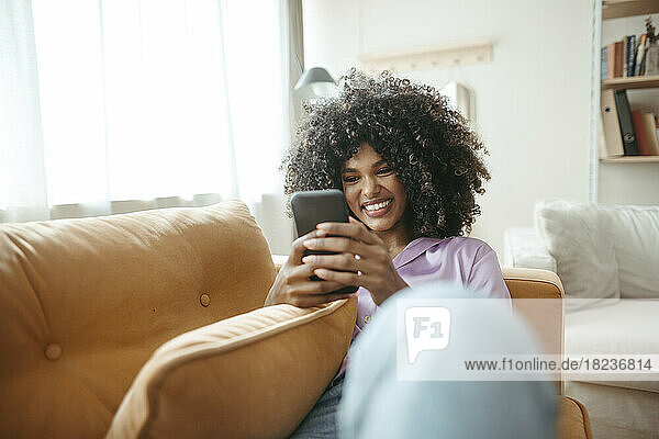 Lächelnde Frau benutzt Smartphone auf Sofa im Wohnzimmer