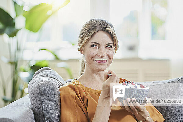 Smiling woman holding yogurt bowl with fruits on sofa at home