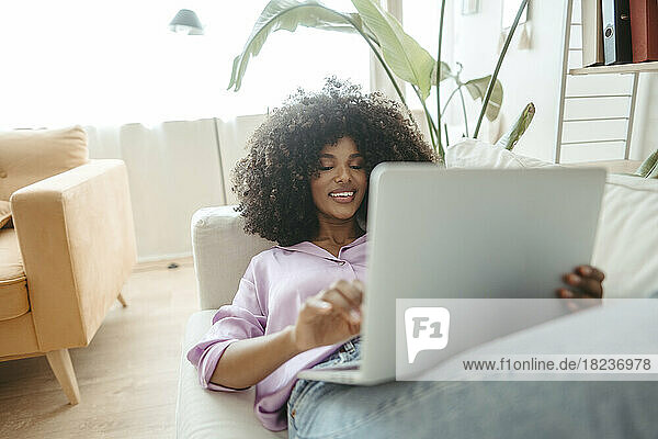 Smiling Afro woman using laptop lying on sofa at home