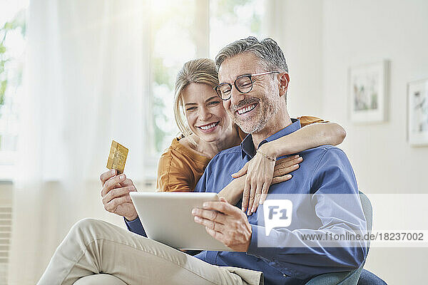 Smiling woman embracing man with credit card doing online shopping through tablet PC at home