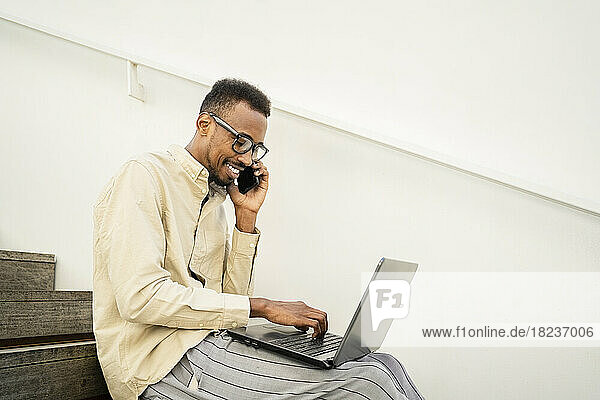 Smiling man talking on smart phone and using laptop by wall at stairs