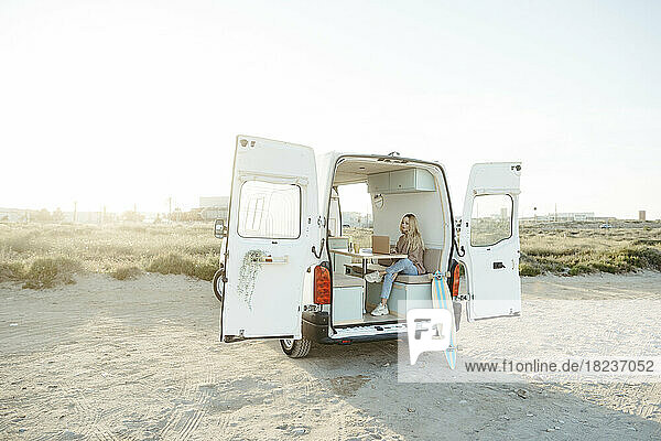 Young freelancer sitting with laptop in caravan in front of clear sky
