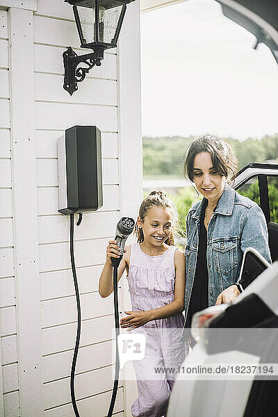 Lächelnde Tochter mit Mutter lädt Elektroauto vor Haus