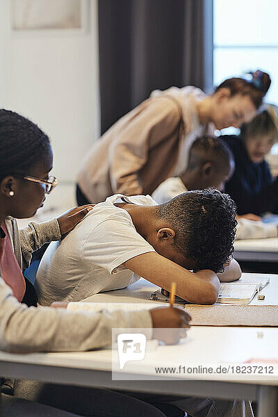 Teenager-Mädchen  das einen deprimierten männlichen Schüler tröstet  der seinen Kopf im Klassenzimmer auf den Schreibtisch legt