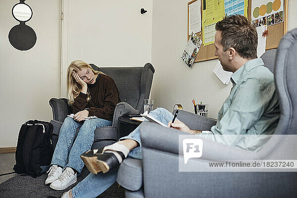 Männlicher Psychotherapeut im Gespräch mit einer depressiven Schülerin  die auf einem Stuhl in einem Schulbüro sitzt