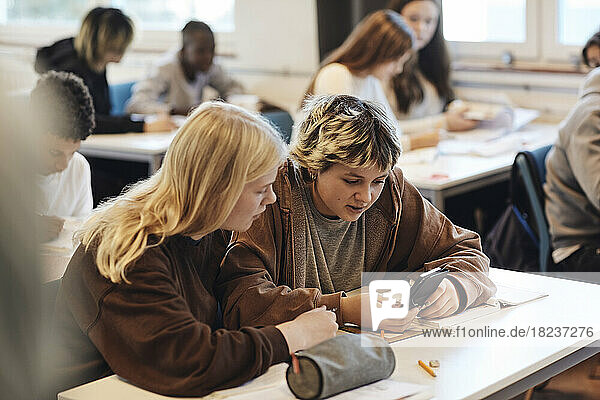 Teenager-Mädchen teilt sich ein Smartphone mit einer blonden Freundin  die am Schreibtisch im Klassenzimmer sitzt
