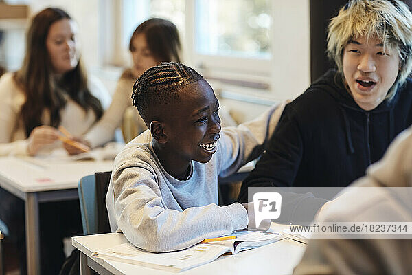 Happy teenage boy sitting by cheerful male friend at desk in classroom