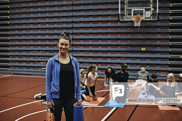 Portrait of smiling female coach standing with exercise mat against students in basketball court