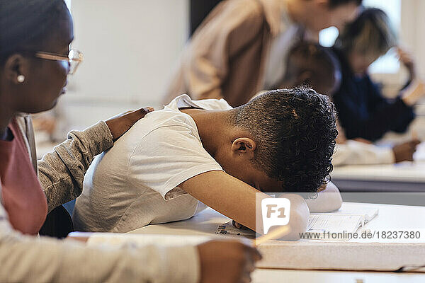 Eine Studentin tröstet einen deprimierten Teenager  der seinen Kopf im Klassenzimmer auf den Schreibtisch legt