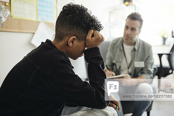 Sad teenage boy sitting by male counselor discussing in school office
