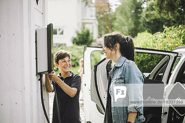 Lächelnder Junge  der ein Kabel hält und seine Mutter in der Nähe eines Elektroautos betrachtet