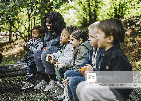 Multiracial boys and girls eating fruits while sitting with female teacher in park