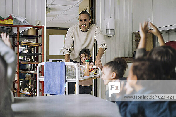 Happy teacher and girl with food trolley by classmates during breakfast in day care center