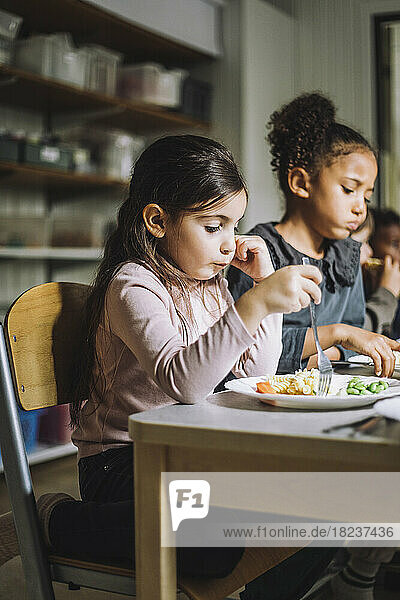 Girl having pasta for breakfast with female classmate in child care center