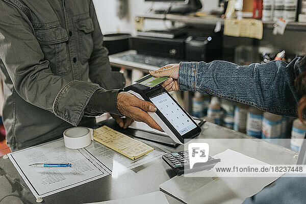 Männlicher Schneider hält ein Kreditkartenlesegerät in der Hand  während eine Kundin in einer Werkstatt kontaktlos bezahlt
