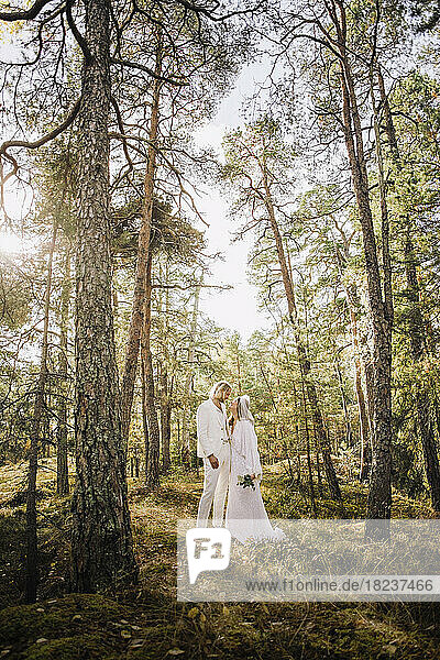Mid adult bride holding bouquet looking at groom standing amidst trees in forest