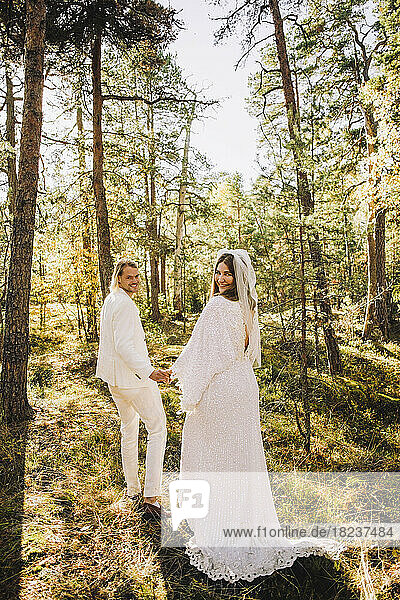 Lächelnde Braut und Bräutigam in voller Länge  die sich an den Händen halten und in den Wald zurückblicken