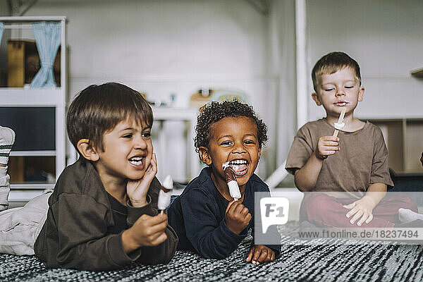 Fröhliche männliche Studenten essen Eis auf dem Teppich in der Kindertagesstätte