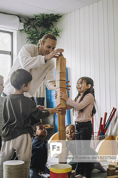 Männlicher Lehrer  der Kinder beim Stapeln von Holzklötzen im Kindergarten anleitet