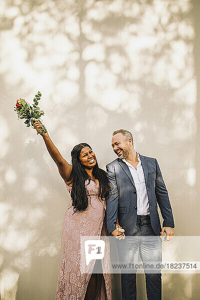 Happy bride holding bouquet with hand raised standing by groom against wall