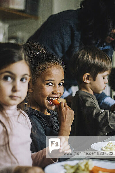 Glückliches Mädchen isst Karotte zum Frühstück mit Klassenkameraden im Kindergarten