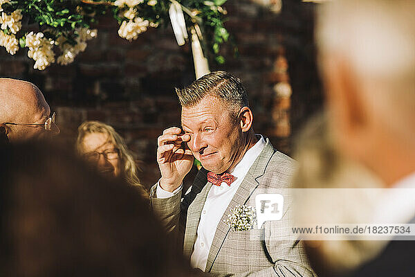 Lächelnder Bräutigam  der sich mit dem Finger die Tränen abwischt  inmitten von Gästen an einem sonnigen Tag