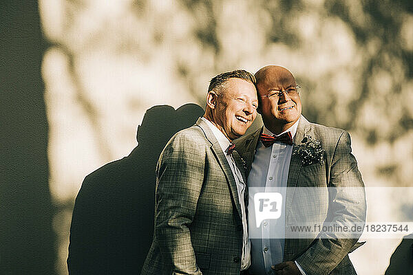 Glückliche gut gekleidete Homosexuell Männer stehen gegen die Wand während der Hochzeit am sonnigen Tag