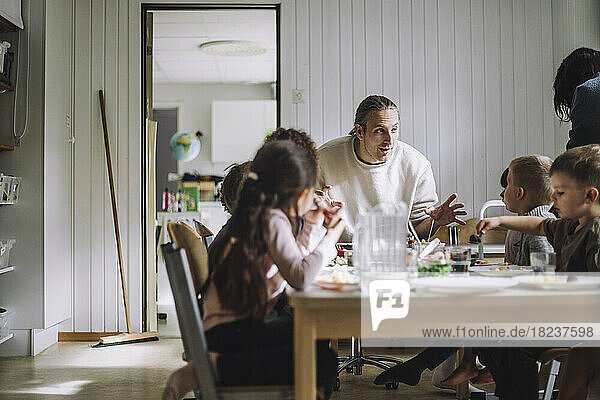 Männlicher Lehrer im Gespräch mit Schülern beim Frühstück am Tisch in einer Kindertagesstätte