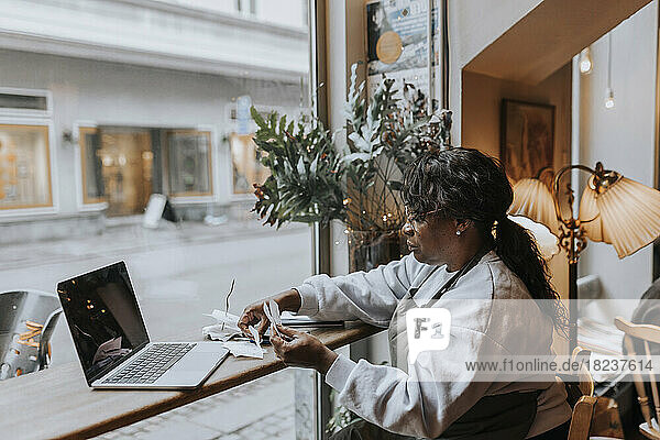 Side view of mature cafe owner with bills and laptop sitting at table by glass window in coffee shop