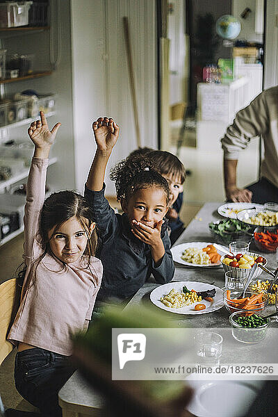 Glückliche Mädchen und Jungen mit erhobenen Händen am Esstisch beim Frühstück im Kindergarten